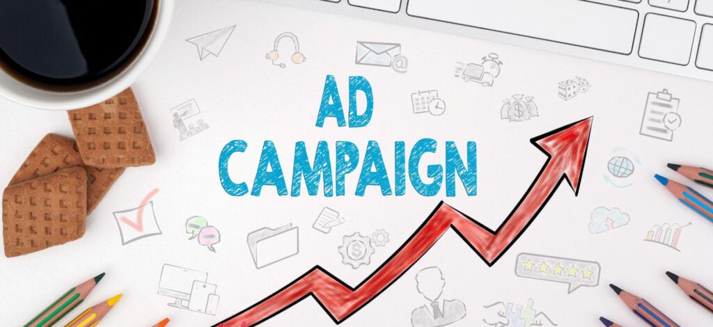 A drawing of a line chart with the word "Ad campaign" in bold, surrounded by small doodles. A keyboard, coffee mug, some biscuits and colored pencils are in the frame.