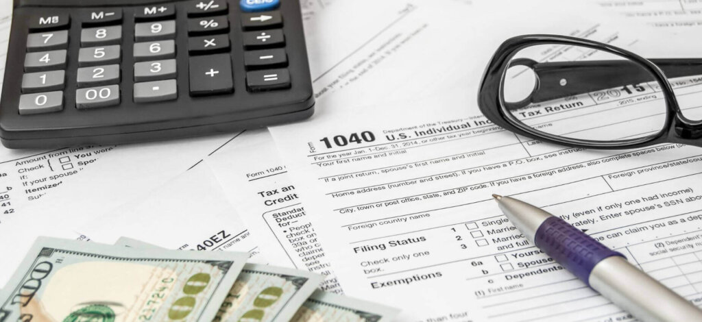 This image features some tax papers sprawled out, a calculator, a pair of glasses, some cash, and a pen.